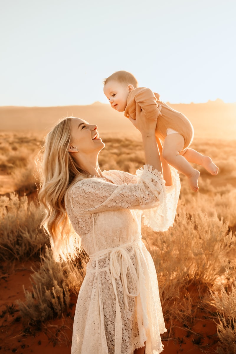 Motherhood Photographer, a happy young mother holds her baby in the air while standing in a field of dry grass