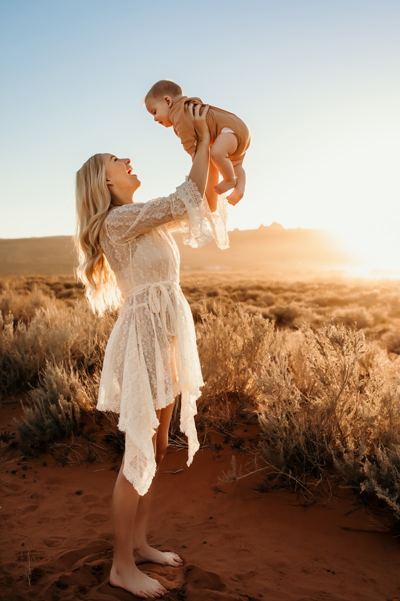 Motherhood Photographer, mother holds baby in the air smiling, they are in the desert