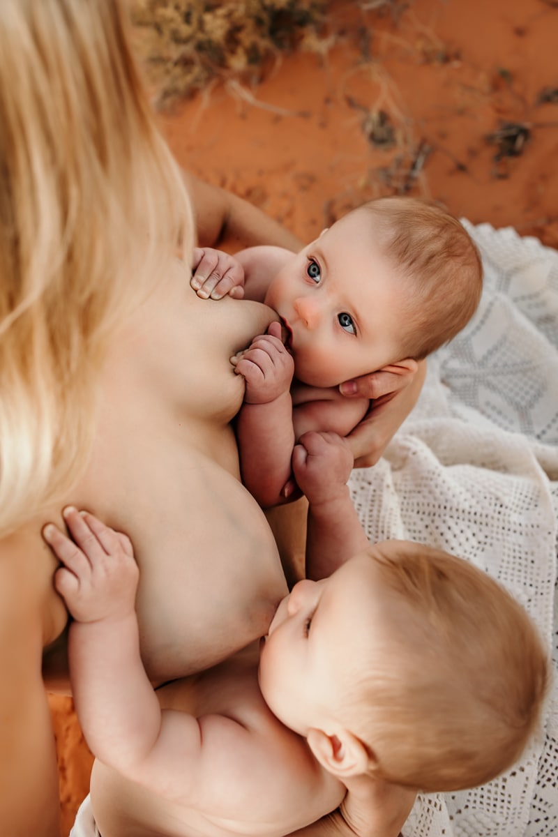 Motherhood Photographer, a mother breastfeeds her twin babies as sits outside in the desert sand
