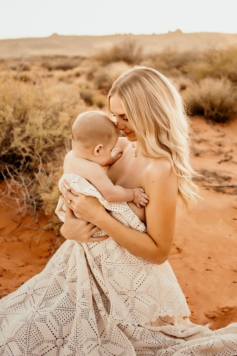 Motherhood Photographer, a young mother holds her baby close as she sits in the desert sand
