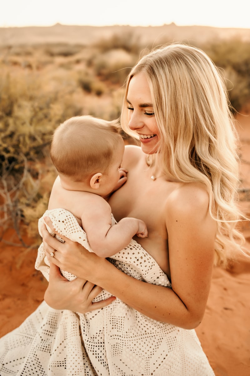 Motherhood Photographer, a young mother holds her baby, she smiles as she stands in the desert sand