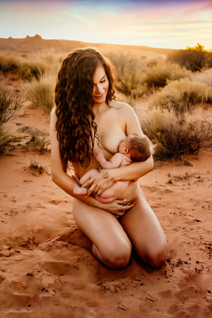 Motherhood Photographer, a nude mother breastfeeds her baby while kneeling in the desert sands