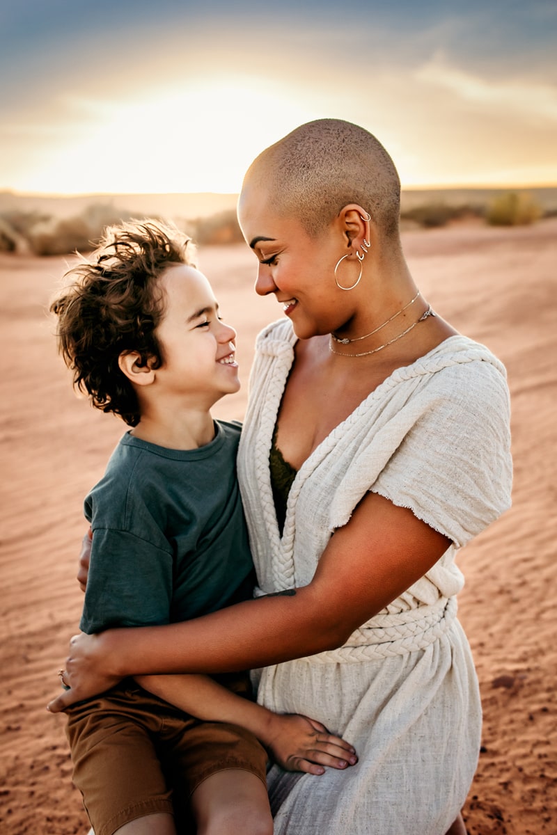 Motherhood Photographer, a mother and son look directly at each other and smile before a setting sun in the desert