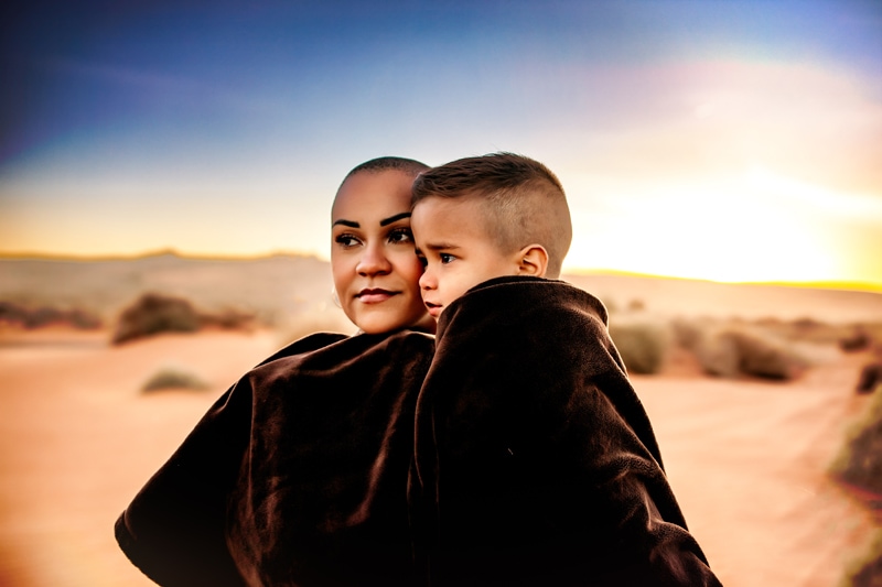 Motherhood Photographer, a mother and her young son are wrapped in a blanket as they stand in the desert at dusk