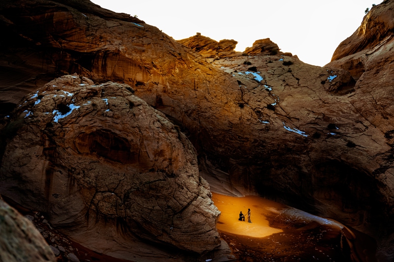 Elopement Photographer, a man kneels before his lover, they are in the sand beneath Utah Canyons