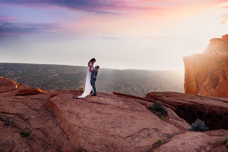 Elopement Photographer, man stands on Utah canyon wall, he lifts his bride in the air