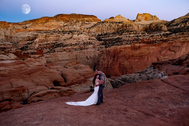 Elopement Photographer, a newly married couple embrace at dusk in the canyons of Utah, the moon full and bright behind them