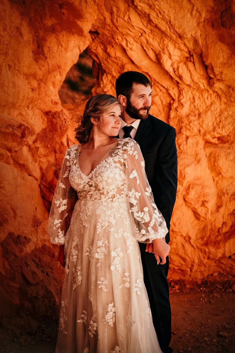 Elopement Photographer, man stands behind woman holding hands before desert wall, she is in wedding dress