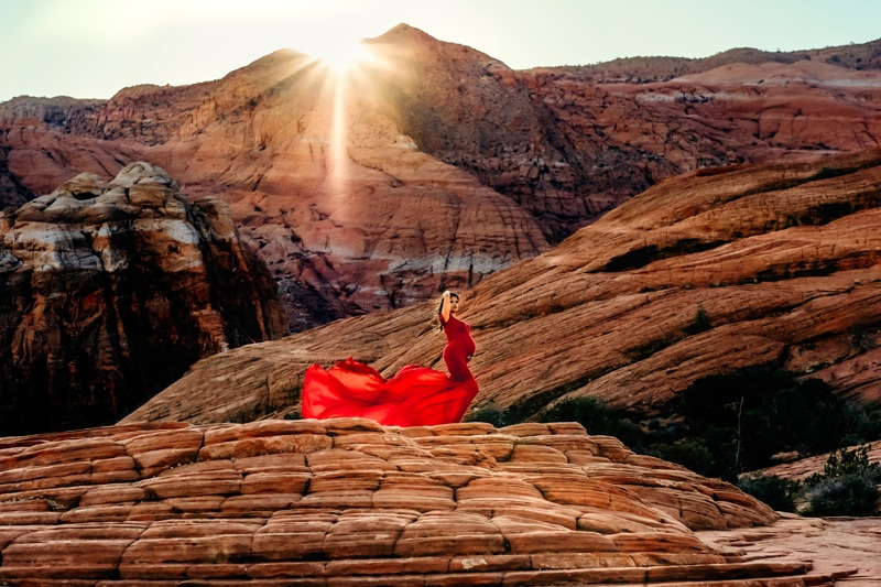 Motherhood Photographer, a pregnant woman wears a long flowing dress as she stands before a desert mountain landscape