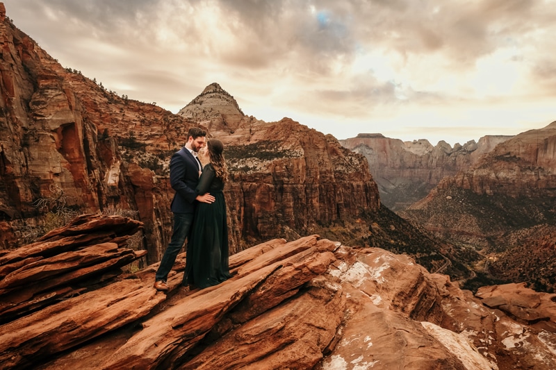 Vacation Photographer, man and woman embrace as they stand on rock above Zion National Park