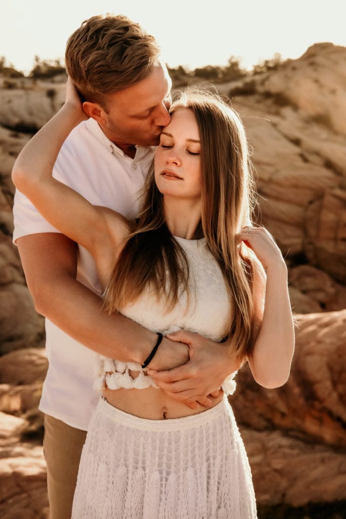 Couples Photographer, Man hugs woman from behind, she embraces him, they are outdoors