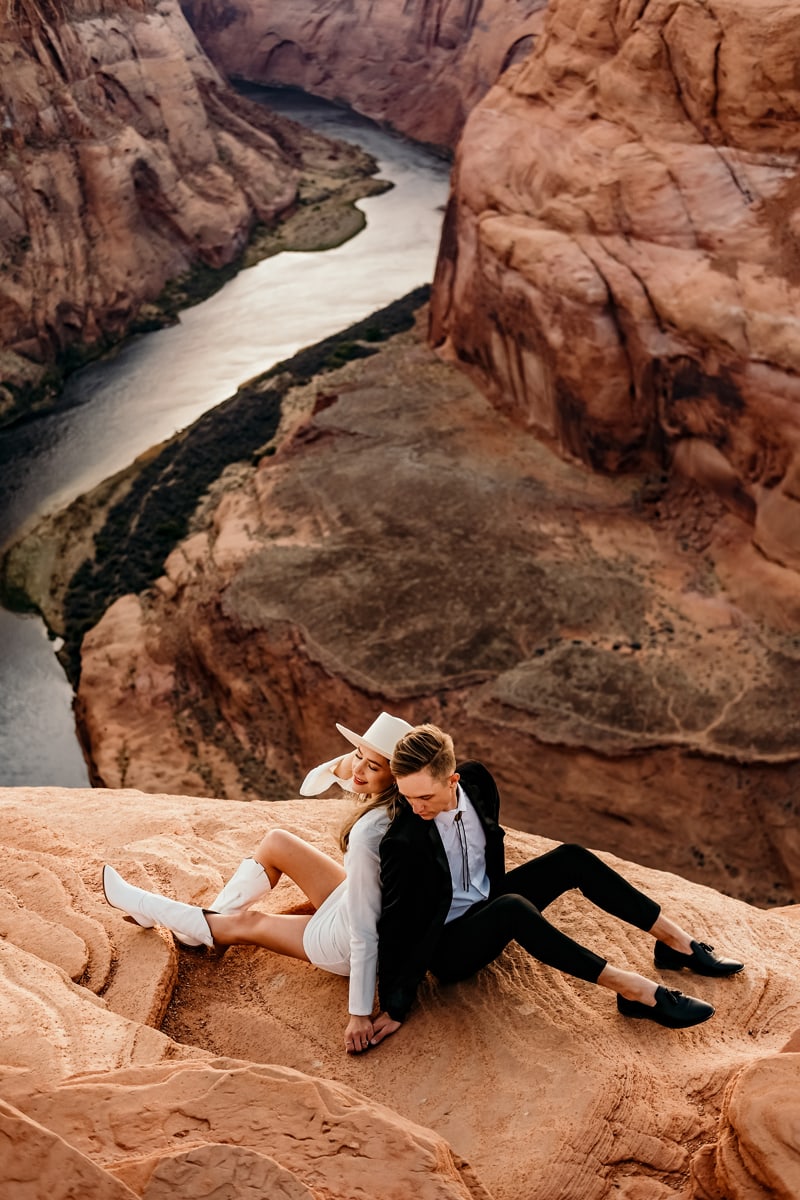 Elopement Photographer, young couple sit back to back, they are freshly married and sit atop a canyon wall before a river