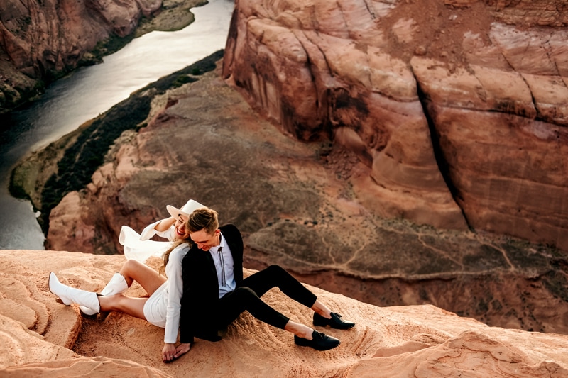 Elopement Photographer, a young married couple sit back to back on the edge of a canyon, the river runs beneath them