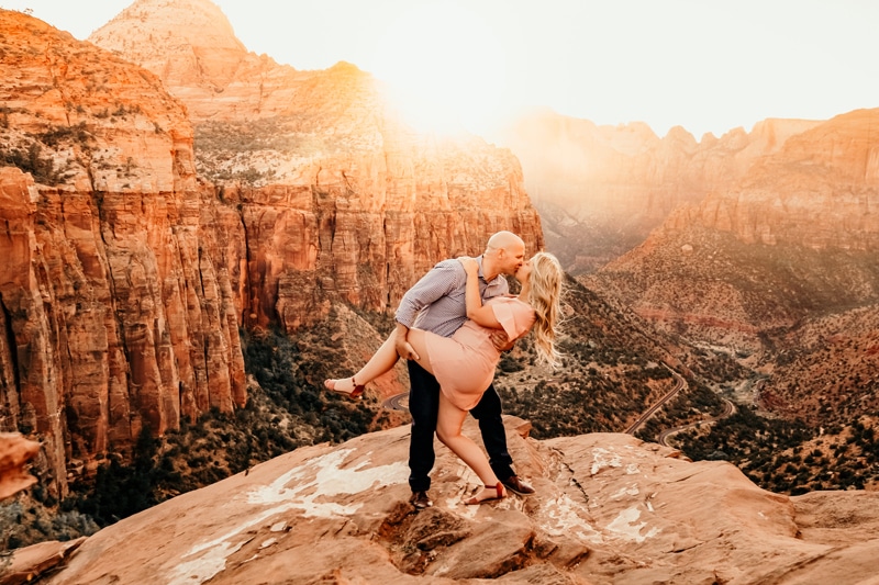 Vacation Photographer, a man and lifts his wife up as he kisses her before Zion National Park
