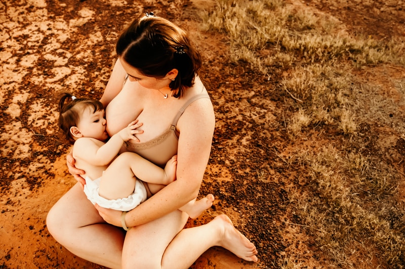 Motherhood Photographer, a woman sits on the ground outside as she breastfeeds her daughter