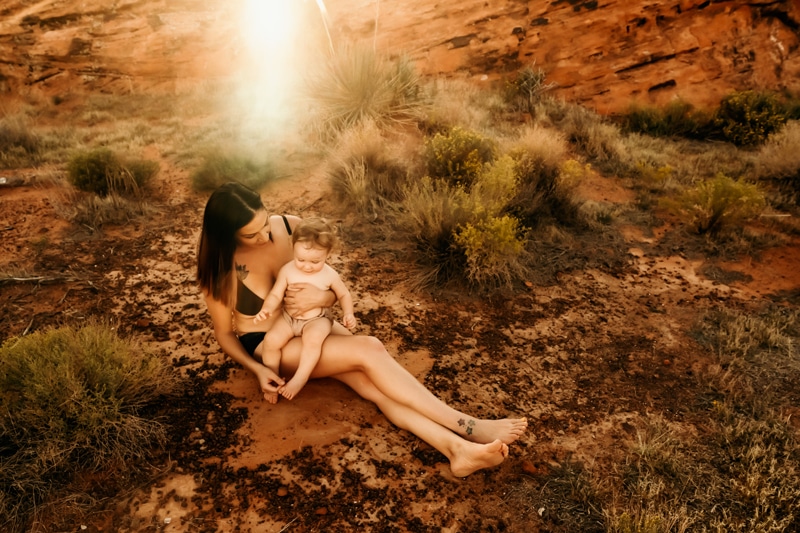 Motherhood Photographer, a mother sits with her baby on her lap near desert shrubs
